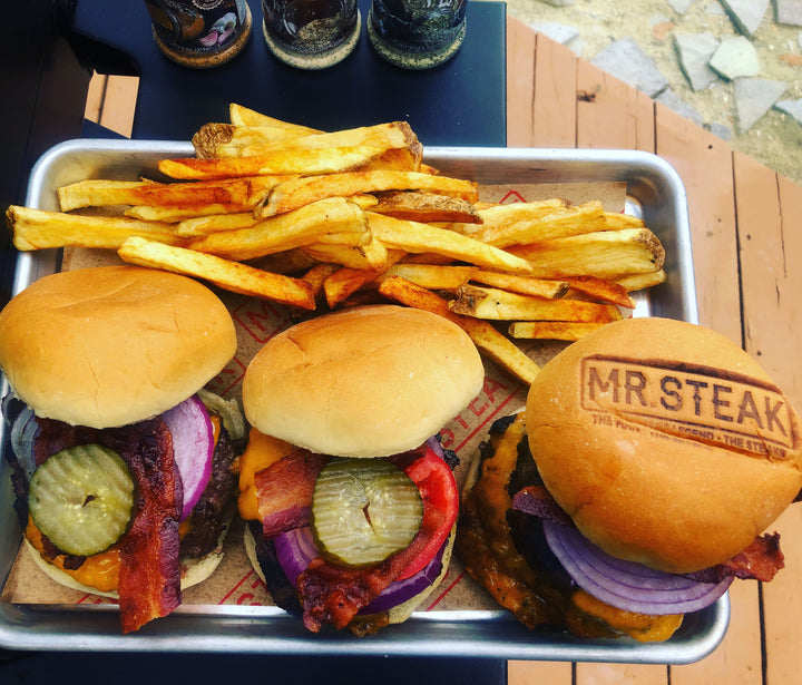 Three wagyu burgers ready to be eaten topped with cheese, pickles, tomato, onion, bacon with a side of fries