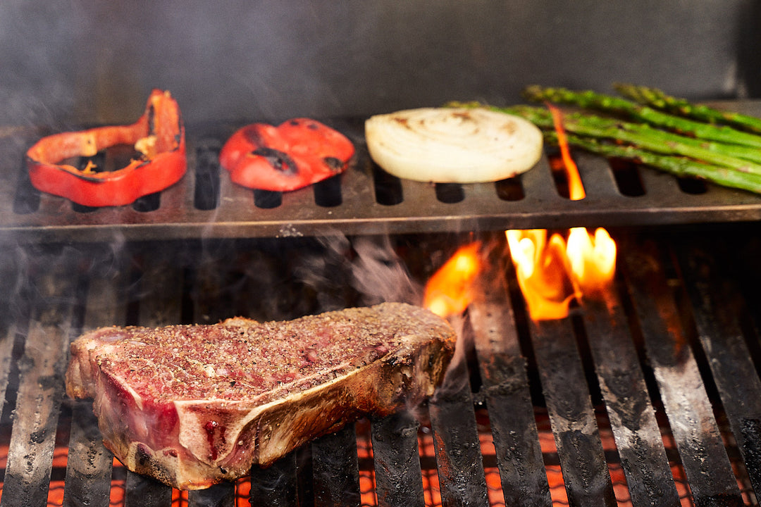 Bone-in New York Strip sizzling on the barbecue along with red peppers, onions and asparagus