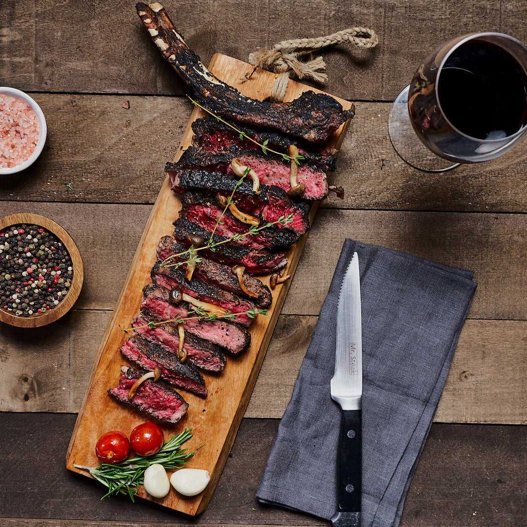 Perfectly cooked tomahawk, cut and plated stunningly with salt, pepper, tomato, rosemary and garlic, sitting beside a Mr. Steak knife